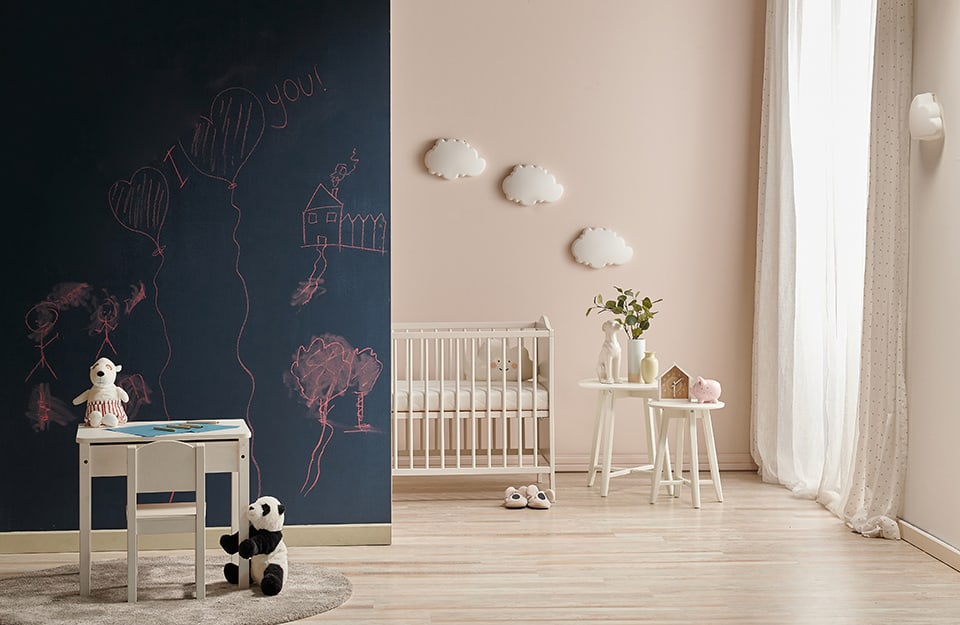 Children's room in an essential style. One wall is painted with chalkboard paint and there are drawings. In front of the wall is a small desk and chair with puppets beside it. Beyond the wall is the rest of the room, painted in pastel pink, with a wooden cot, small tables and cloud-shaped wall decorations;