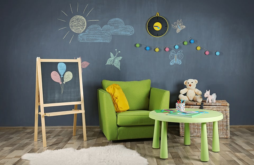 An area of a children's room with a wall painted with chalk paint and drawn on. In front is a green armchair, a small green plastic table and a blackboard with three coloured balloons drawn on it in chalk;