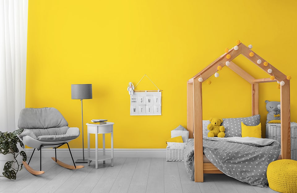 Child's bedroom with yellow walls, a wooden canopy bed in the shape of a small house, with yellow and white pompoms hanging from it, grey parquet flooring, wicker baskets, grey and yellow puppets, a grey rocking chair, with a small table next to it and a standing lamp;
