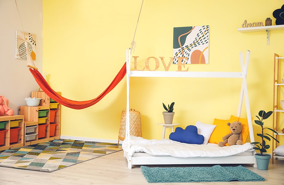 Children's bedroom with a bed with a wooden curtain frame, a light yellow wall, shelves with storage units like drawers, a red hammock hanging on the wall, wooden signs around the room, puppets and a carpet with triangular geometries;