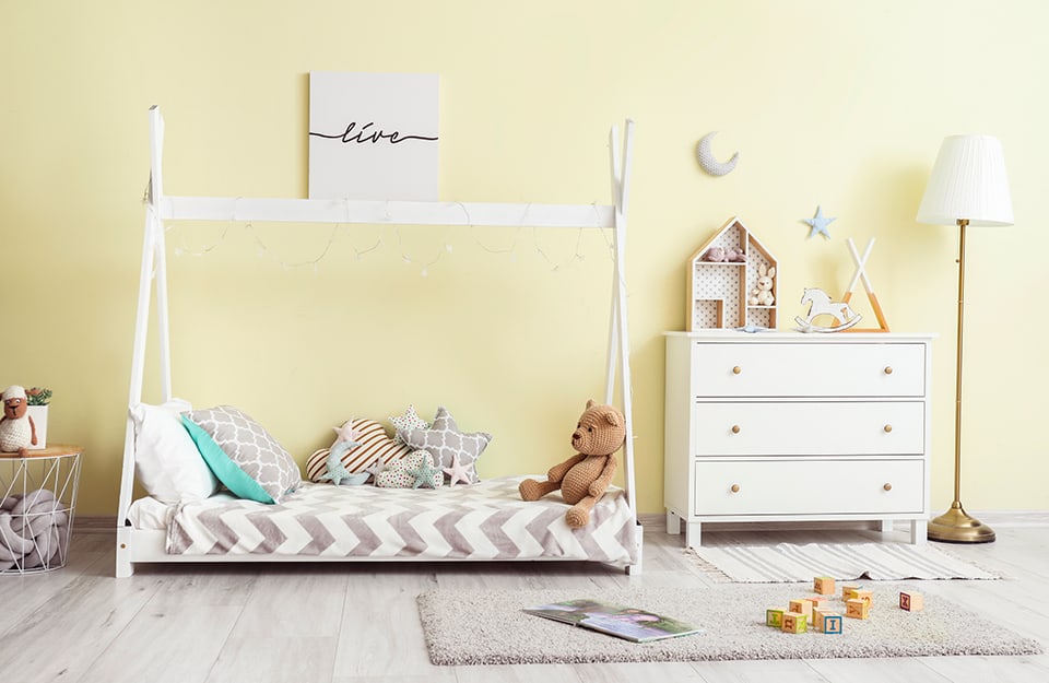 Children's room with light yellow wall, white wooden bed with curtain frame, white chest of drawers, carpet, grey parquet floor and various accessories and toys scattered around the room