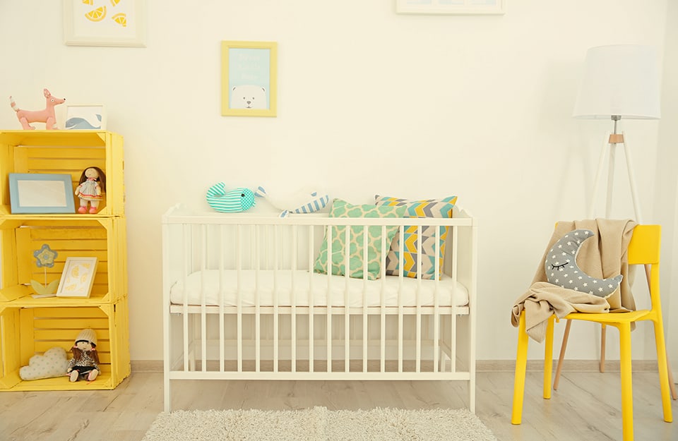 Baby's room with soft yellow walls, white cot, yellow chair, yellow shelf made from fruit crates and several hanging pictures;