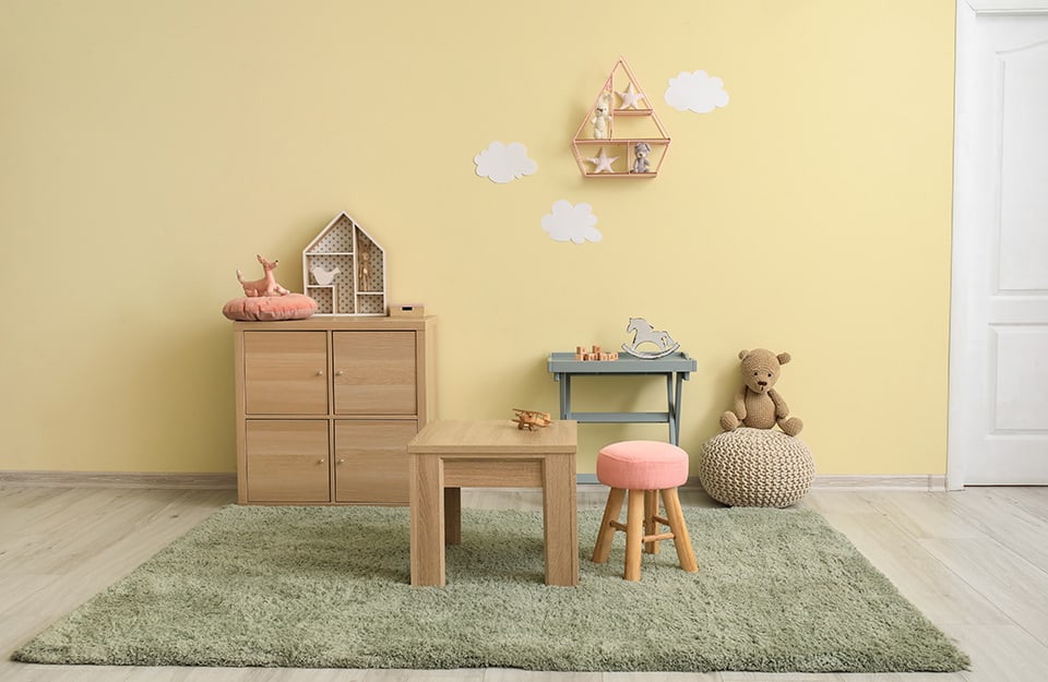 Children's room with a small wooden table and a stool with a pink seat above a green carpet. On the yellow wall are a wooden shelf, a small work table and a puff with a puppet on it;