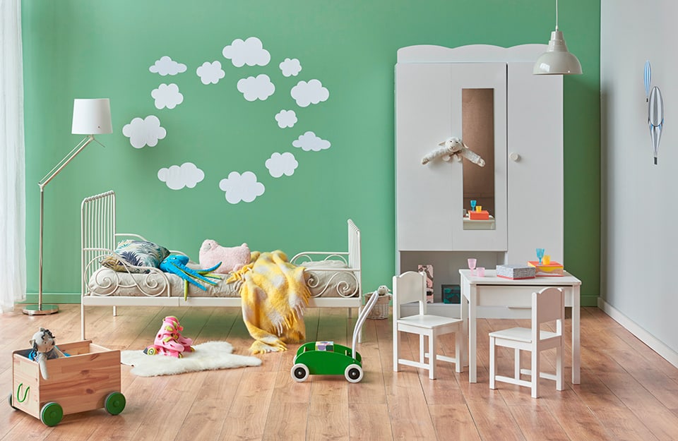 Children's bedroom with parquet flooring and a green wall with white cloud decorations. The wardrobe is white, with a mirrored door, and the bed is also white, with a metal frame. There is a white floor lamp and, as well as toys on the floor, a small white table with two small chairs, also white;