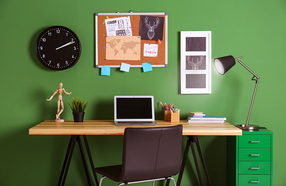 Children's room desk, with a green wall with a clock on it, a cork board with various things attached and a print with a drawing of a deer's head. On the desk is a small drawing dummy, an open laptop computer, a floor plan, filled pencil cases, blocks and notebooks. Next to the desk is a deep green chest of drawers;