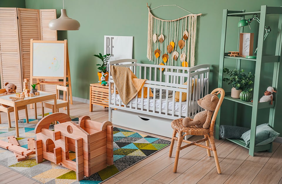 Children's bedroom with parqut, green wall, pure green bookshelf, white cot, wooden tables and chairs, a marker board and, above a geometrically patterned carpet, a structure made of wooden constructions