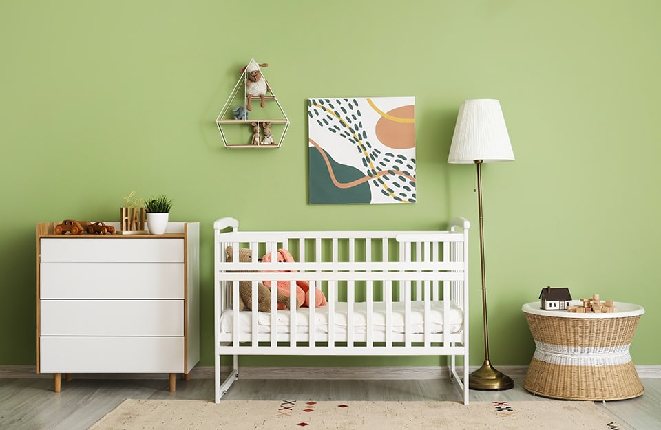 Dormitorio infantil con cuna y cómoda blancas, lámpara blanca, mesa de mimbre y pared verde, con una estantería de formas geométricas angulosas y un estampado abstracto en tonos pastel colgado;
