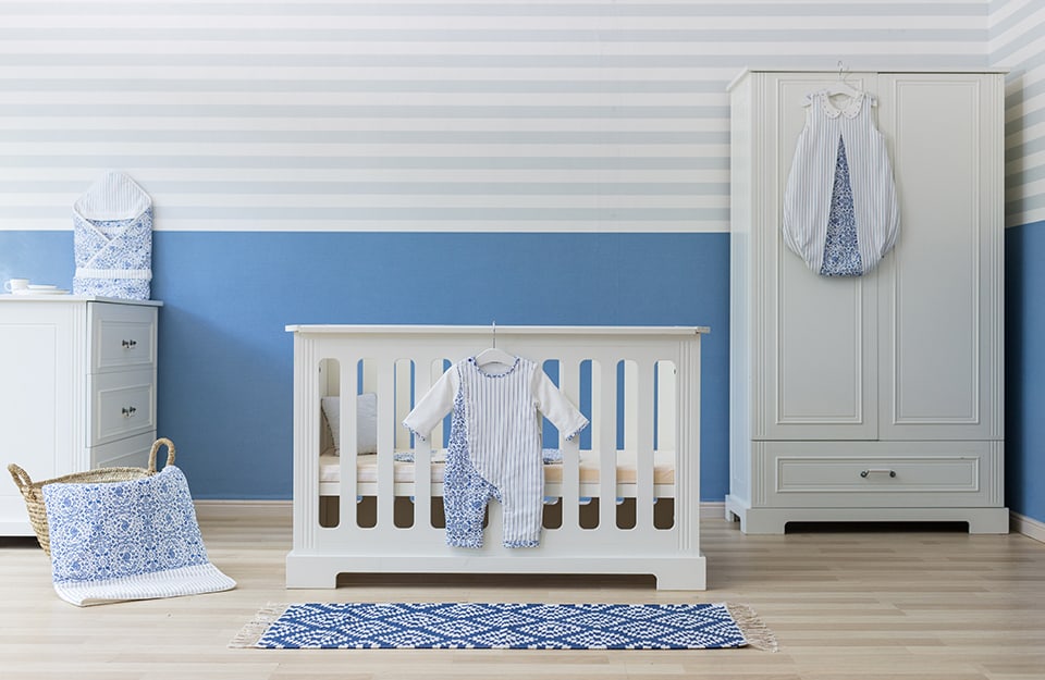 Dormitorio infantil con cuna blanca clásica, armario blanco y ropa de colgar blanca y azul. El suelo es de parqué y la pared está pintada de azul suave en la parte inferior y de rayas horizontales azules y blancas muy suaves en la superior;