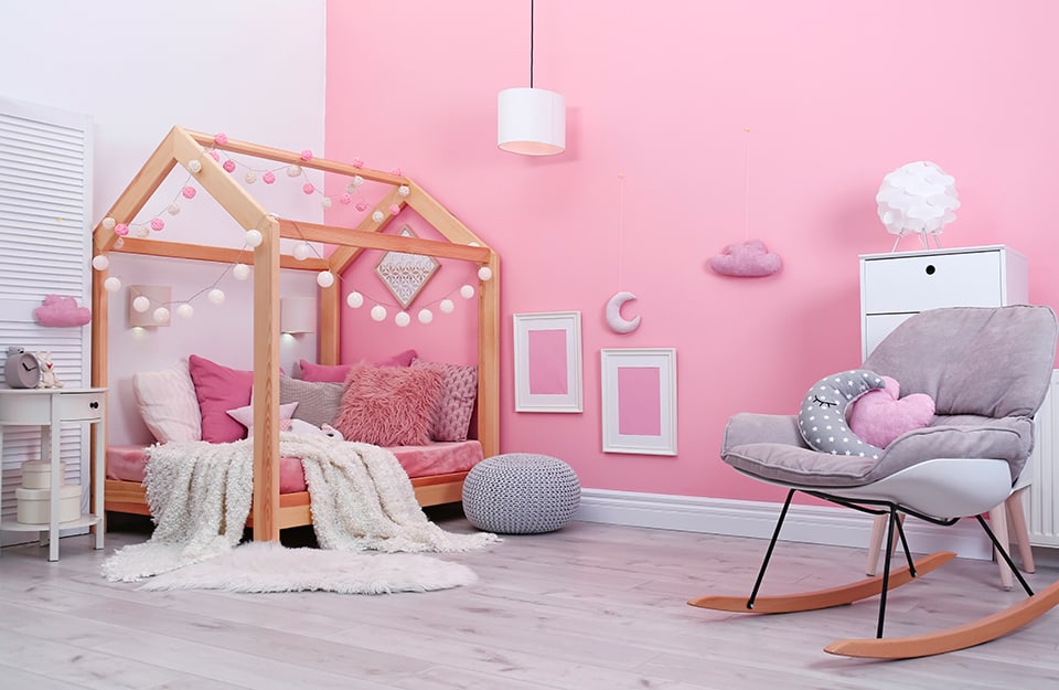 Cameretta per bambini con parete rosa decorata con quadri, una luna e una nuvoletta di tessuto, letto a baldacchino con struttura in legno a forma di casa, decorata con pompon bianchi e rosa, tanti cuscini rosa, una poltrona a dondolo grigia con cuscini