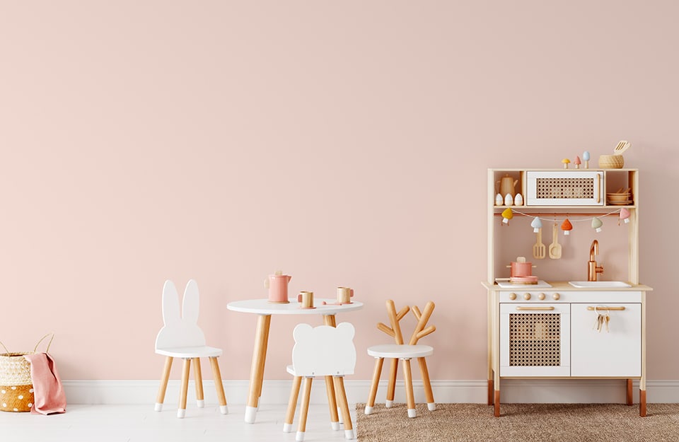 Pastel pink wall of a children's room, with small wooden toy kitchen and small table and chairs in shades of natural wood and white;