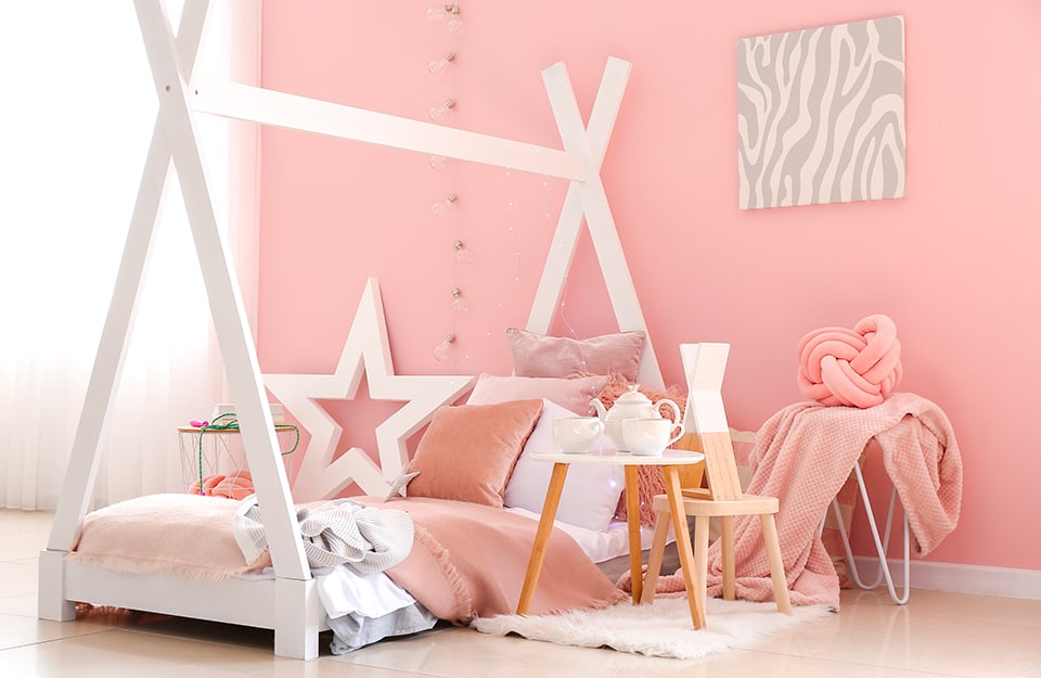 A pink bedroom with a bed with a white wooden frame in the shape of a curtain, bed linen in shades of pink, a pink wall with a row of light bulbs descending from the ceiling, a grey abstract painting and wooden coffee tables, one of which has a tea service on top;