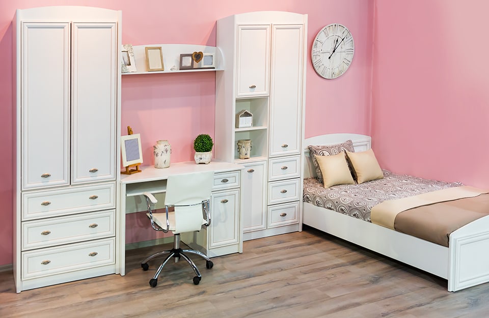A bedroom with pink walls, a bed with a wall clock hanging on it and a double white wardrobe with a desk in the middle;