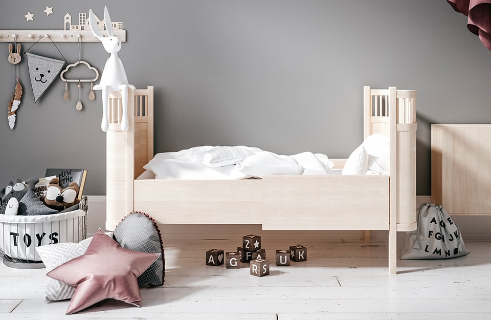 Wooden cot bedroom with an unusual shape, under which you can see toy cubes with letters printed on them. The floor is a vintage white parquet. The wall is grey, with a coat rack decorated with the outline of a city on it. There are cushions on the floor and a basket with 'TOYS' written on it and toys inside;