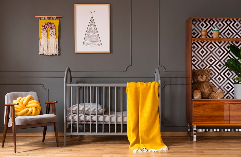 Room in shades of grey and yellow with a dark grey wall with frames in the same colour, prints on the walls, a wardrobe without doors with an interior upholstered with geometrically patterned paper, a grey cot with a yellow blanket, a grey Scandinavian-style chair with a yellow woven cushion;