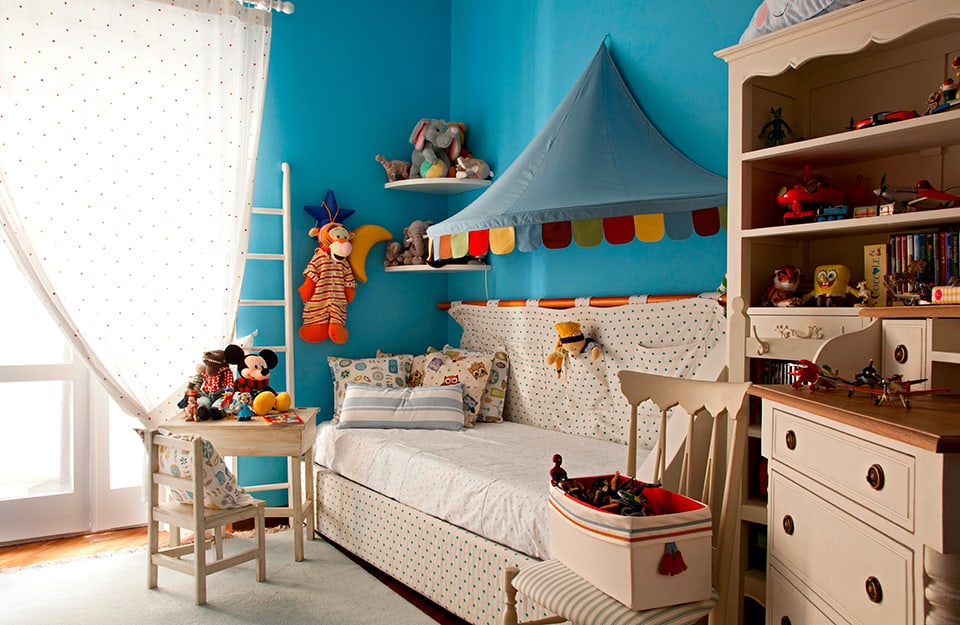 Sofá cama y modernos muebles de madera de estilo vintage en una habitación infantil muy colorida y juguetona. Encima del sofá hay una especie de carpa de circo y la pared es azul celeste