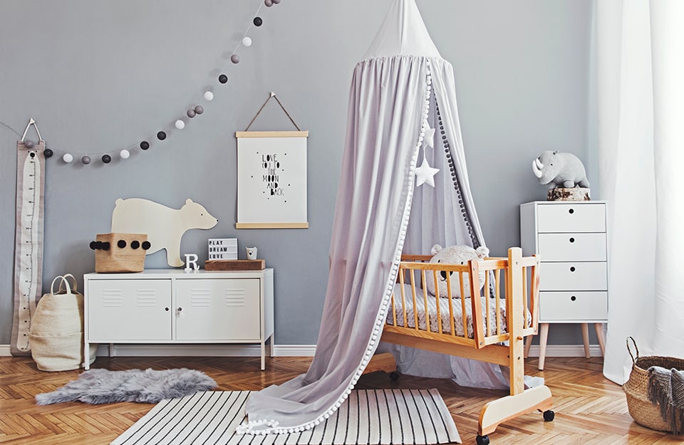 Children's room with wooden cradle and grey canopy. The wall is also grey, with white, grey and black pompoms hanging from it. The floor is parquet. There are white cabinets and several scattered toys;