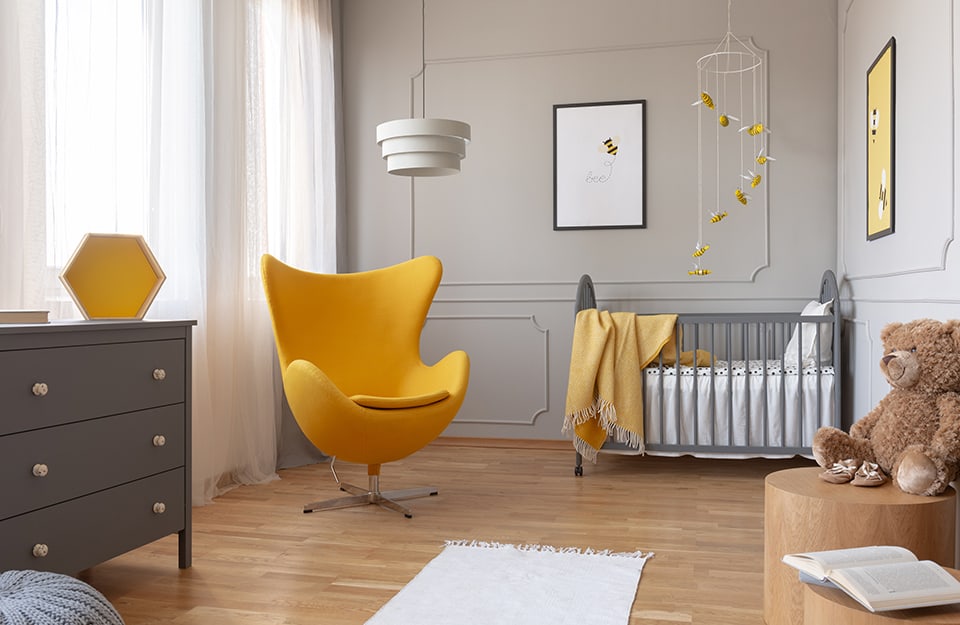 Cemera children's room in shades of grey and yellow, with grey wall, grey cot, grey chest of drawers, parquet floor, yellow designer armchair, white chandelier, on the walls prints with bees, and bees above the cot as toys;