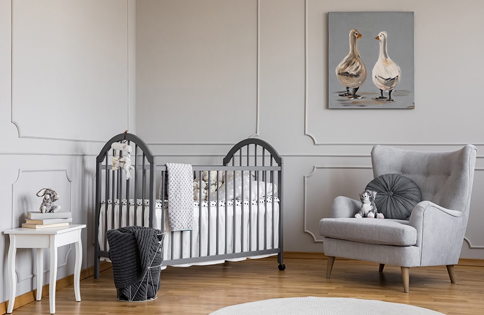 Children's bedroom with a dark grey cot and white bed linen. The wall is grey, decorated with frames of the same colour. The floor is parquet. There is a large grey armchair, a white circular carpet, a small white table and a picture with two painted geese;