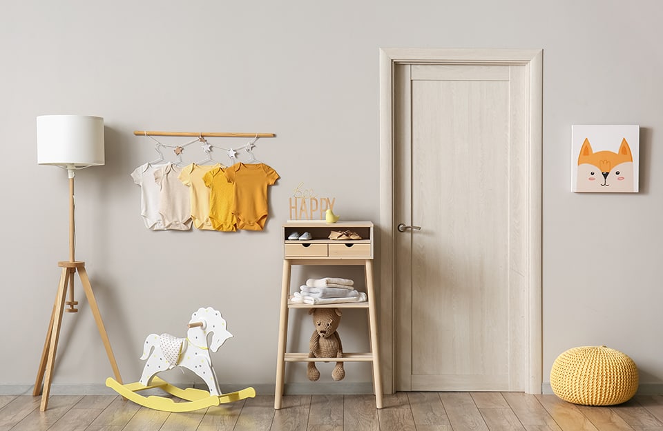 The wall of a small bedroom, in light grey, with a door, a wooden towel rack, a wall hanger with a baby bodysuit in shades of yellow, a white and yellow rocking horse, a white lamp with a wooden stand and a fox face print on the wall;