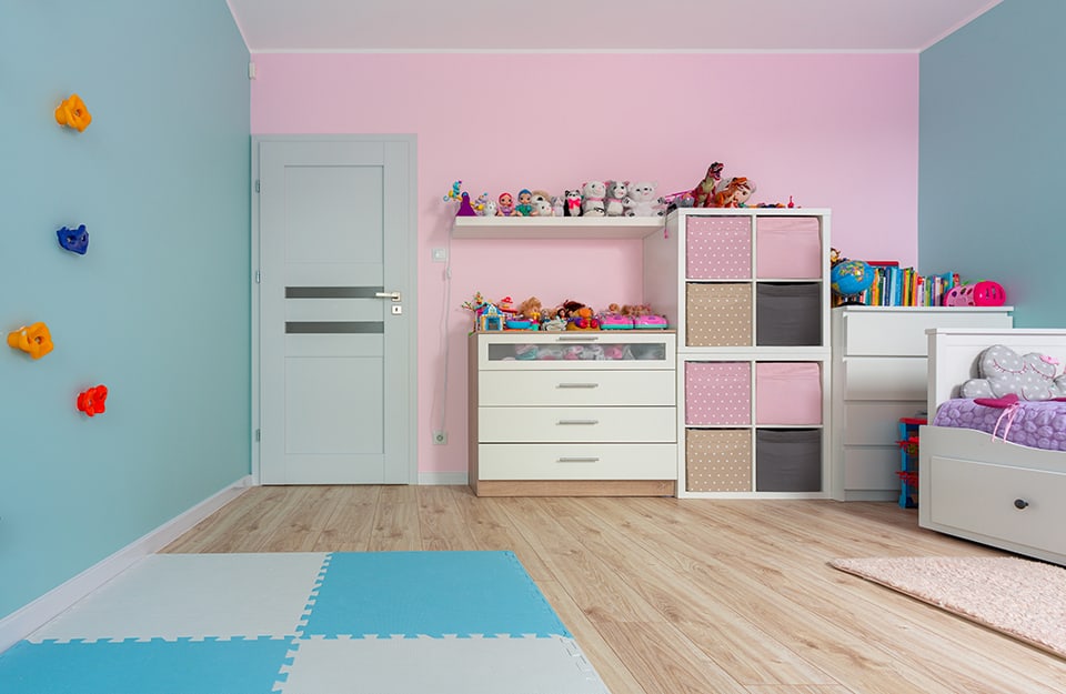 Children's room with pink and blue walls, furniture with fabric drawers in different colours and patterns, white chest of drawers, shelves with lots of coloured dolls on them, light blue play mat, white bed with purple blanket and indoor free climbing wall;