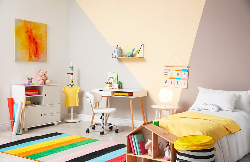Room with a grey wall decorated with a painted yellow triangle. The carpet is multi-coloured with stripes, as are the other elements in the room, including a bed, a desk, a small shelf and a low hanging rail;