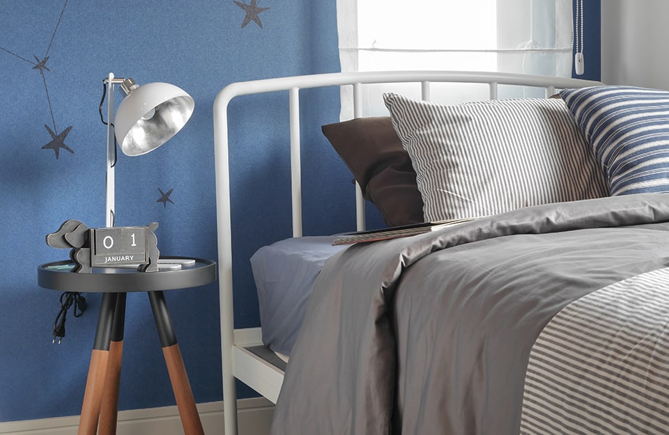 Detalle de un dormitorio infantil con cama de metal blanco, pared azul, mesilla de noche trípode con lámpara encima y calendario en forma de perro. En la pared hay dibujadas constelaciones en negro