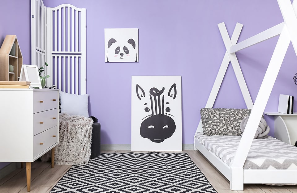 Bedroom with lilac wall with two prints (one depicting a panda and the other a zebra), a black and white geometric-patterned carpet, a white bed with a curtain frame and black and white geometric-patterned bed linen, a white chest of drawers and a screen also in white;