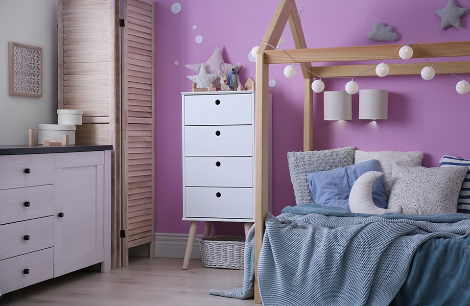 Children's room with white wall and lilac wall. The bed is made of wood with a house-shaped frame, there are several white cabinets and decorations. The linen is on blue and light blue