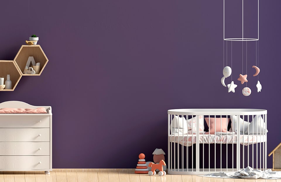 Intense purple wall of a children's bedroom, with white cot, carousel with planets and stars, changing table with drawers, hexagonal shelves and parquet floor