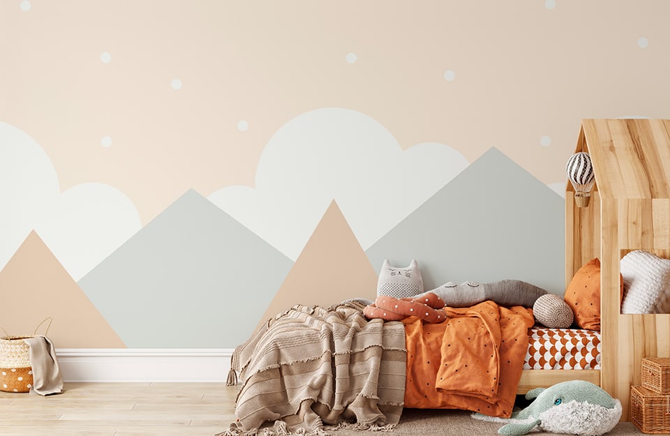 Children's bedroom with mountain-themed wall decoration in shades of pink, white and pastel blue. There is a wooden bed with a cottage-style headboard. Above the bed several blankets and pillows