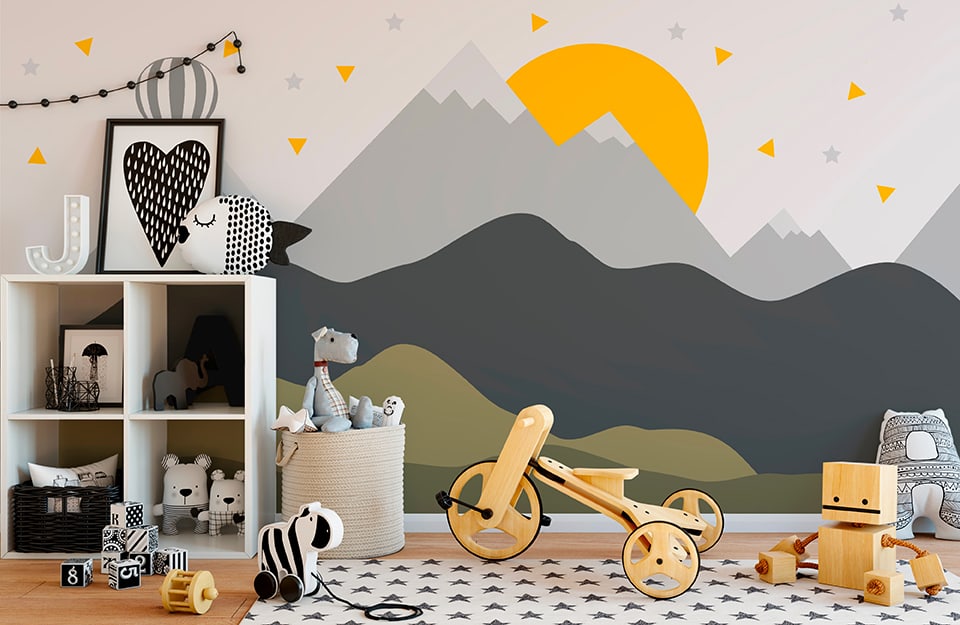 Children's bedroom with mountain-themed wall decoration in shades of yellow, grey, white and green. There is a white shelf with toys, objects and moulds all in black and white. The floor is parquet, with a white carpet with black stars. There is a basket full of puppets and there are several toys on the floor, including a wooden and rope robot and a wooden tricycle