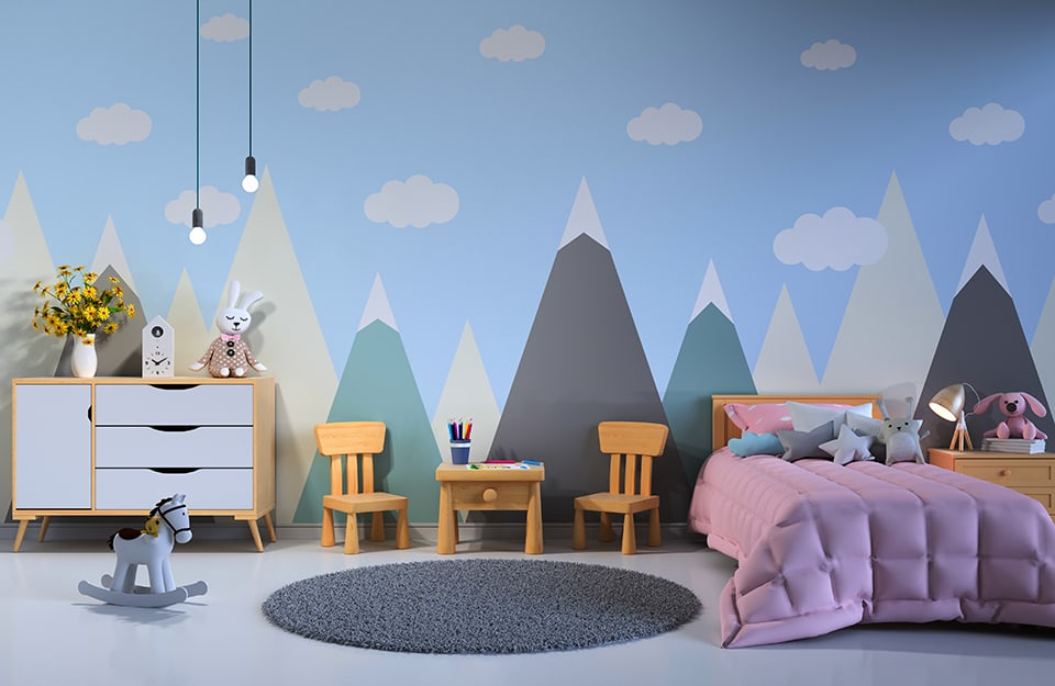 Children's bedroom with mountain-themed wall decoration in shades of blue, white, grey and green, with a bed with a purple blanket, a grey circular carpet, a wooden bedside table with a puppet and a bedside lamp, a small wooden table with two small chairs, a white cupboard and a mini rocking horse on the floor. Two light bulbs descend from above;