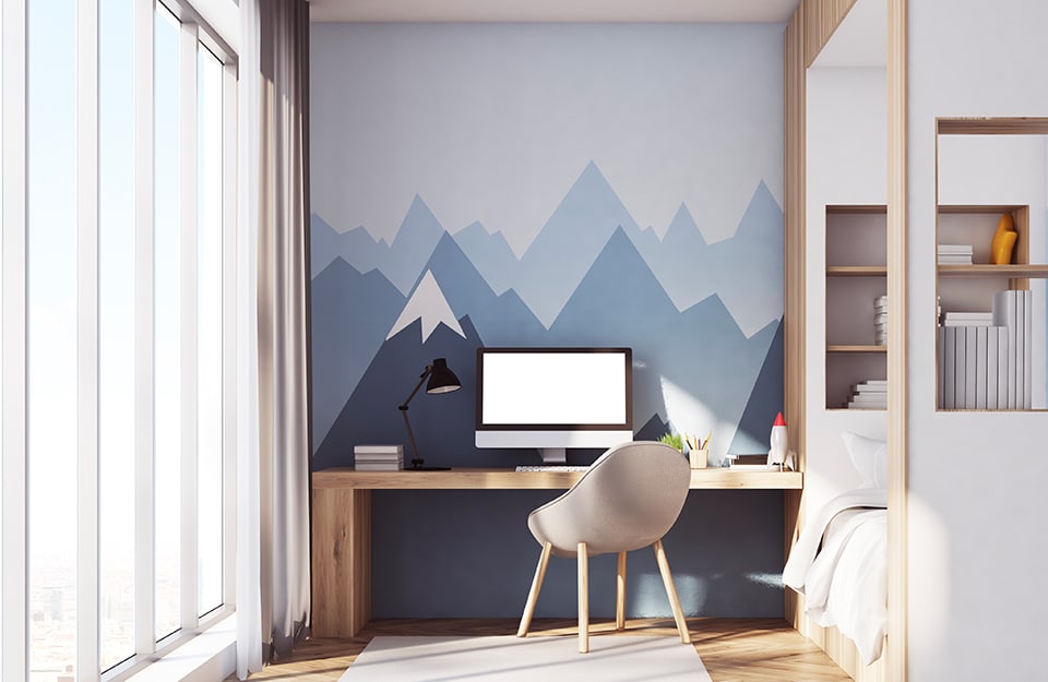 A boys' bedroom with a mountain-themed wall decoration in shades of light blue and blue. There is a large wooden desk with a computer on it, a work lamp, books and a toy rocket. The chair is modern. To the right is a bed inside a natural wood and white painted wood frame that also serves as a shelf;
