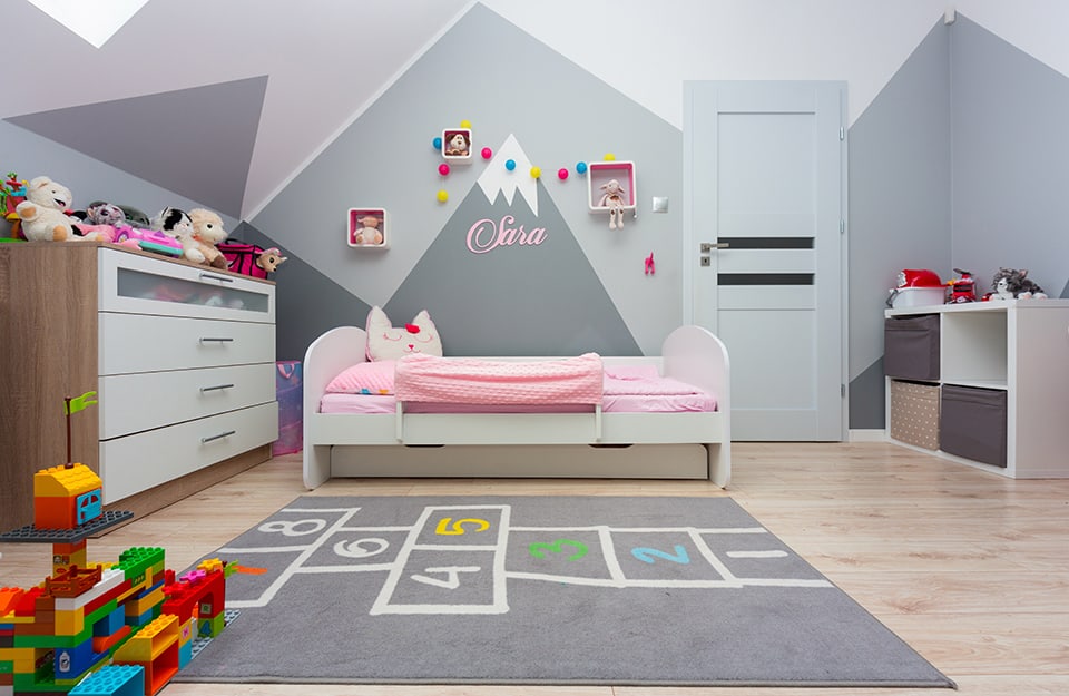 A little girl's bedroom with mountain-themed walls decorated in shades of white and grey. There is a bed with pink bedding and a cat-shaped pillow, a grey carpet with bell game fencing on it, a white chest of drawers, a white shelf, shelves with toys and Lego constructions on the floor;