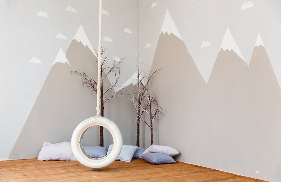 Corner of a children's room with a wall decorated with a mountain theme in shades of white and grey. In the corner are cushions and real tree branches. A white rope hangs from the ceiling with a white-painted tyre-tree hanging from it;