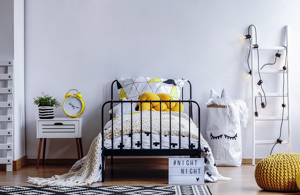 A minimalist children's bedroom in shades of white, with a white wall, white furniture, a black metal bed, black and white bed linen, as well as the geometrically patterned carpet, on parquet flooring. The accent colour is yellow, which we find on the large alarm clock above the bedside table, on one of the cushions, and on an ottoman. In front of the bed is a sign saying 'NIGHT NIGHT' and a heart. Next to the bed is a ladder that acts as a shelf, with lights on it;