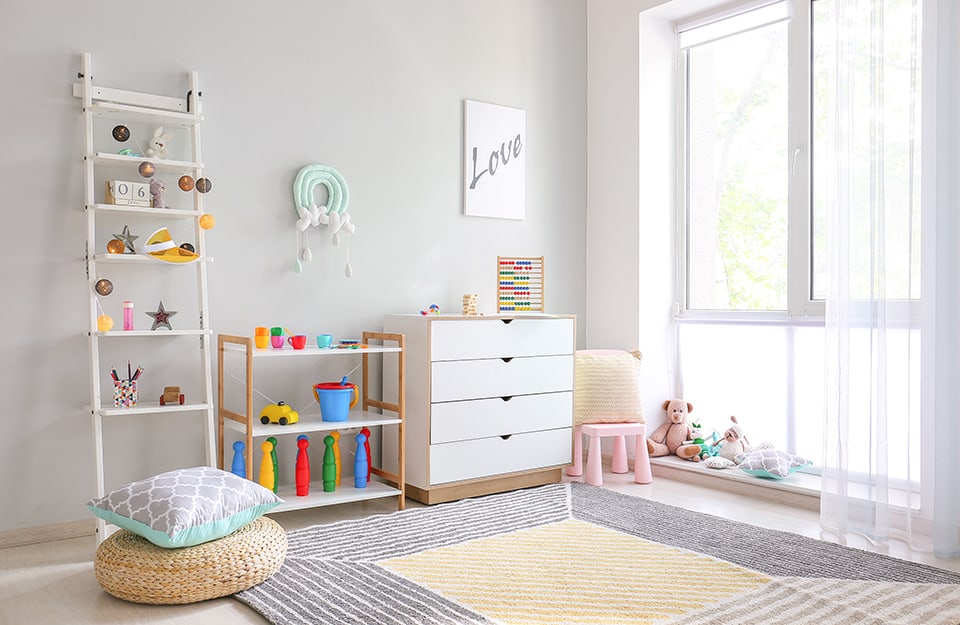 A corner of a children's room in shades of white, with a bright window, a chest of drawers with toys on it and a print saying 'Love', a shelf with more toys, a ladder-shelf leaning against the wall with many knick-knacks and puppets under the window. On the floor is a white floor and a carpet with white, grey and yellow geometric patterns;