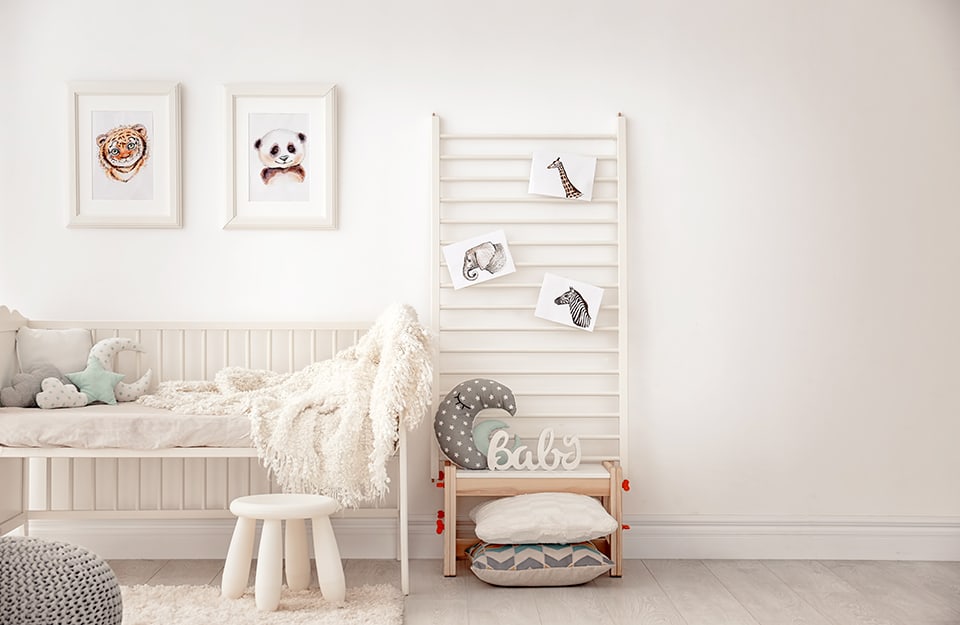 Children's room in total white, with a cot without a side rail, a stool, a small table with cushions, a ladder-shaped shelf leaning against the wall with drawings of animals from the savannah and prints hanging on the wall with other animals;