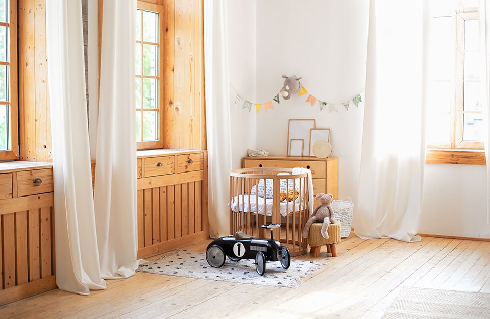 Corner of a children's room in Scandinavian style in shades of white and natural wood. The windows are made of wood set into a wood paneling with drawers. The floor is parquet and above it is a small white carpet with black dots and a toy car that serves as a tricycle. The other furniture is made of natural wood