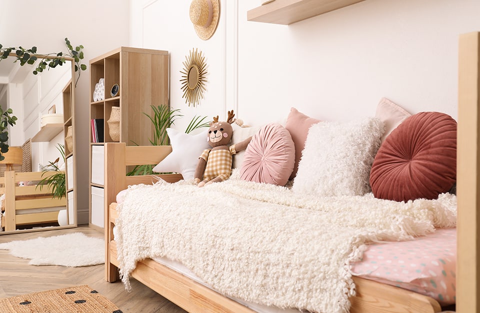 View from the corner of a Scandinavian-style children's room in shades of white and natural wood. The bed is filled with pillows and a shelf can be glimpsed above. Hanging on the wall is a straw hat and a sunburst mirror. Next to it is a wardrobe and further on a mirror that reflects the rest of the room;