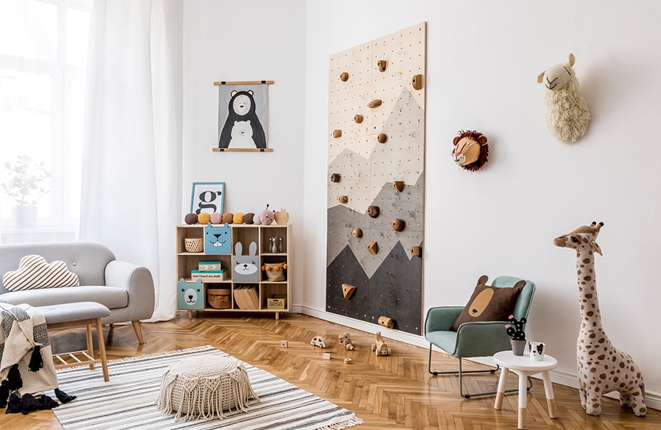Scandinavian-style children's bedroom, with grey sofa, indoor free climbing wall, designer furniture including seats and shelves, herringbone parquet floor, black and white horizontally striped carpet, several puppets around the room and hanging on the wall