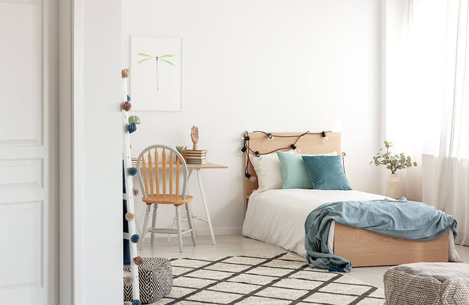 I overlook a Scandinavian-style children's room in shades of white and natural wood. There is a single bed with light blue and green coloured pillows, a desk with books and knick-knacks on it, a print with a dragonfly drawn on it, a large black and white diamond carpet