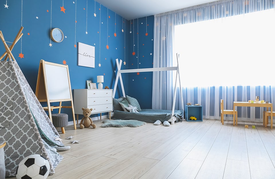 Cameretta per bambini con pareti blu decorate con elementi colorati, letto con struttura a tenda, cassettiera bianca, pavimento a parquet, lavagnetta e giocattoli sparsi