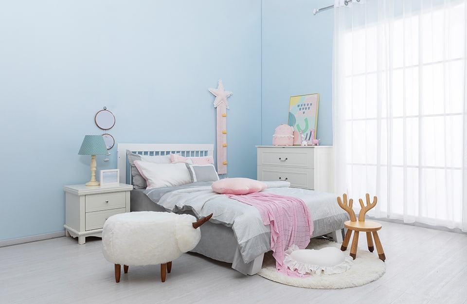 Children's bedroom with blue walls, white bed, large bright window, light-coloured parquet floor, white bedside table and chest of drawers, small chair with backrest in the shape of a deer antler and small hairy table with antlers in the shape of a baby goat;