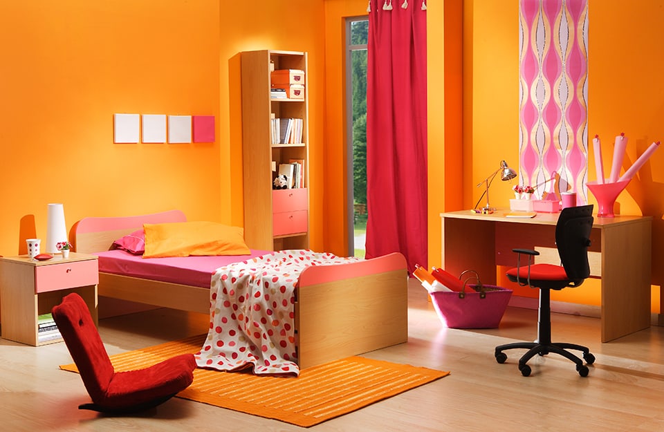 Children's bedroom all in shades of orange and red, with orange walls, cushions and carpet, red chair and curtain, and wooden desk, bed, bedside table and bookcase;