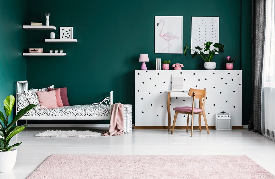 Little girl's bedroom with deep green walls, white floor, pink carpet, bed with metal frame and white sheet with black polka dots. Above the bed are white corner shelves with knick-knacks. At the end of the bed is a white plasterboard structure with black polka dots, with more knick-knacks and two prints on it. In front there is a small table and a chair, as well as a small amplifier. Almost all the decorative elements in the room are in white or pink;