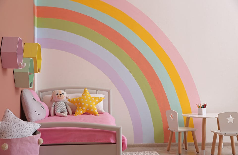 Room with a pink wall decorated with a large coloured rainbow. There is a bed with pink linen, a small table with small chairs, and hexagonal shelves coloured in yellow, pink and green;
