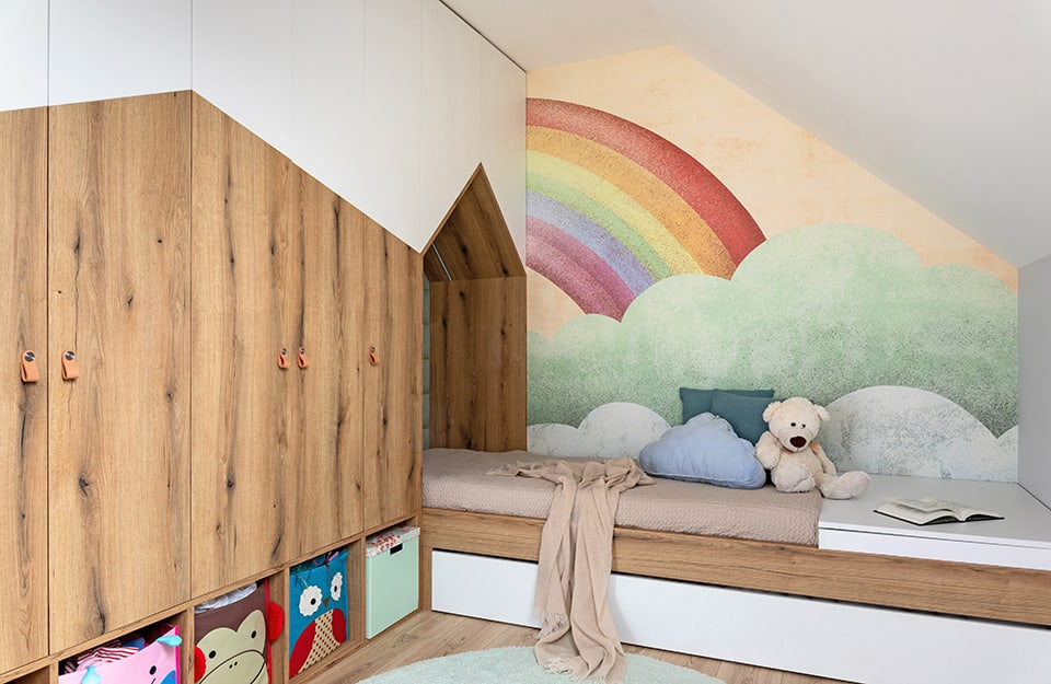 Bedroom with wardrobe-bed system with special white and natural wood doors with broken lines. The bed enters a kind of gallery with a cottage roof and the wall behind the bed is decorated with the design of a cloud with a rainbow