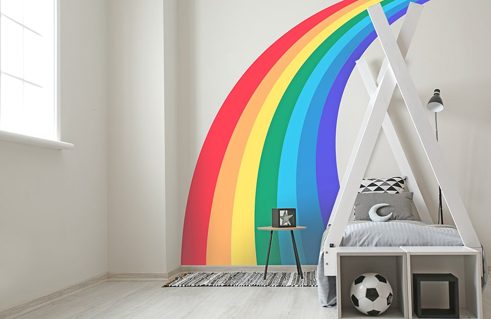 Children's bedroom all in shades of white, with a wall decorated with a rainbow that passes over a bed with a wooden curtain frame. In front of the bed are two floor shelves and in one is a football;