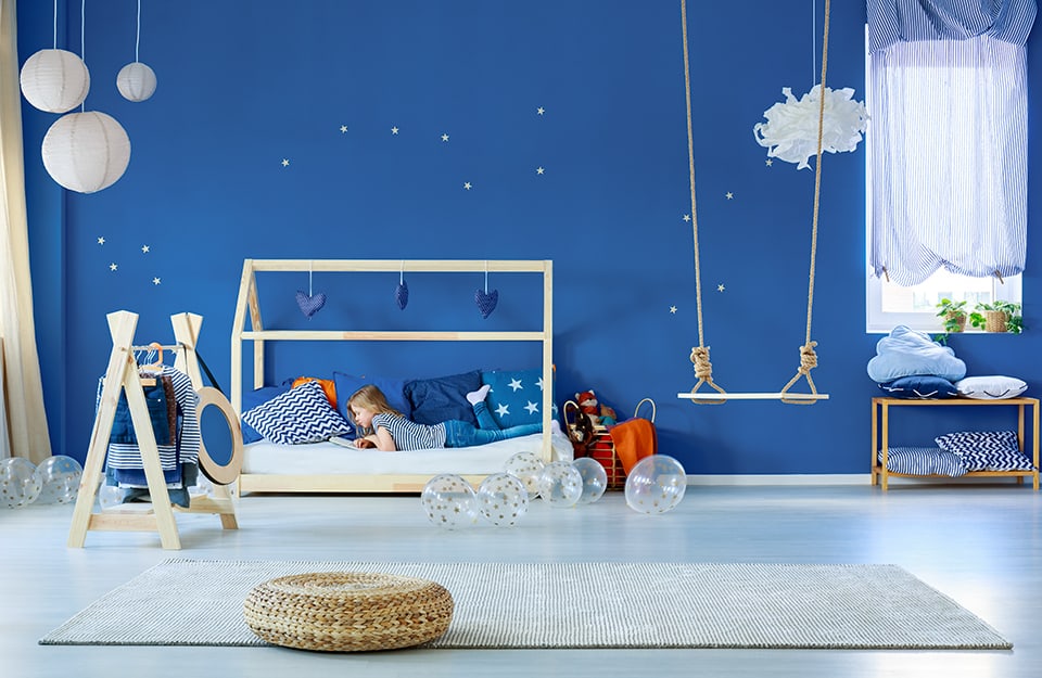 Large bedroom with blue walls. On the bed, with a cottage-like structure, is a blond girl reading. Three spherical chandeliers made of white rice paper and a rope swing descend from the ceiling. On the wall are little white stars. There is a window with blue and white striped curtains. On the floor are transparent balloons with printed golden stars, and a light-coloured carpet. Next to the bed is a coat rack in the shape of a tepee. Under the window is a small wooden shelf with cushions;